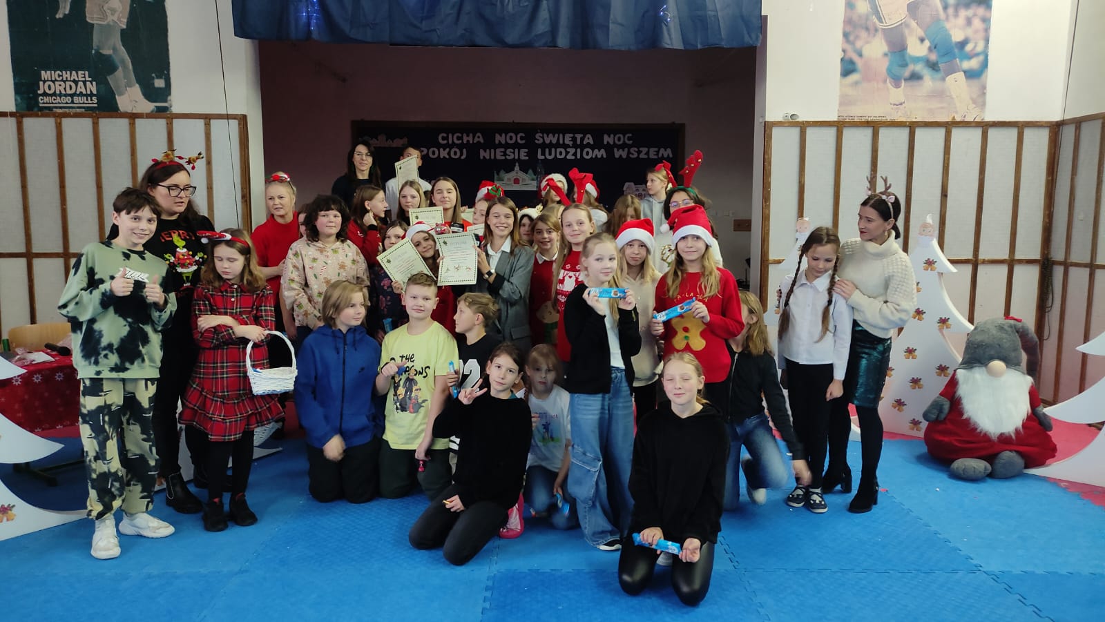 Grupa uczniów stojąca w sali gimnastycznej i pokazująca dyplomy.Niektórzy uczniowie mają na głowach czerwone mikołajowe czapki.