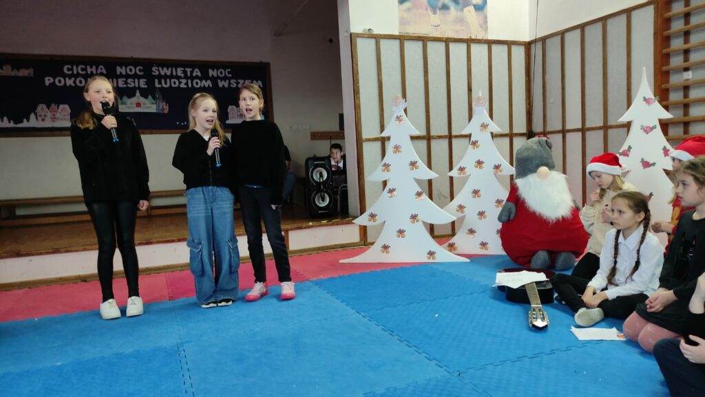 Po prawo trzy dziewczynki trzymaj膮ce mikrofony i 艣pewaj膮ce. Po lewej stronie dekoracje- trzy styropianowe choinki, i tr贸jka dzieci siedz膮ca na pod艂odze i patrz膮ca na wyst臋puj膮ce dziewczynki.