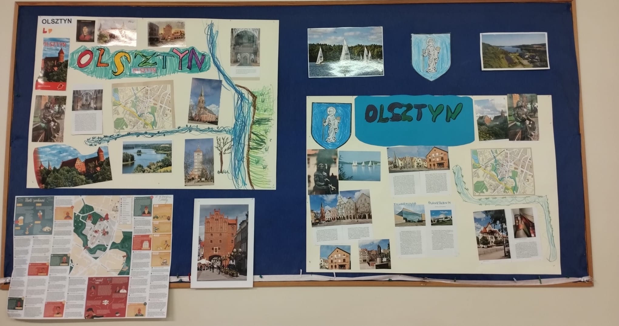 Na zdjęciu widzimy gazetkę ścienną. Na granatowym tle wiszą dwie prace plastyczne wykonane przez dzieci. Prace te to na dużym papierze pokazane zabytki i znane postacie Olsztyna. Dzieci namalowały także rzekę Łynę oraz jezioro. Na gazetce wywieszone ilustracje jezior , herb Olsztyna, zdjęcie Wysokiej Bramy, mapa miasta.