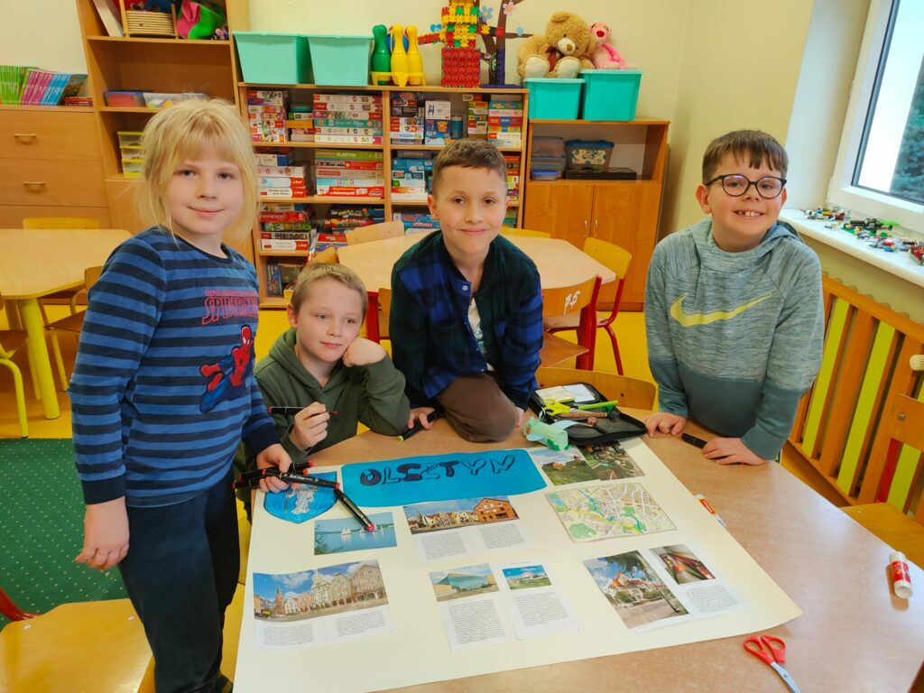 Na zdjęciu widzimy salę świetlicy. Czworo chłopców stoi przy brązowym stoliku. Chłopcy ubrani są w kolorowe ubrania, jeden z nich jest w okularach. Na stole lezy duży , beżowy papier, na którym dzieci ponaklejały zdjęcia i wiadomości o Olsztynie. Na drugim planie widać stoły a dalej regały z kolorowymi zabawkami.