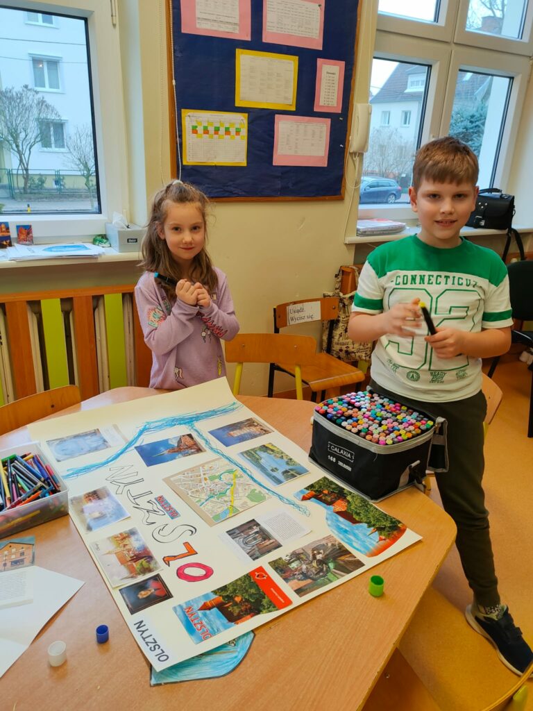 Na zdjęciu chłopiec i dziewczynka stoją przy brązowym, drewnianym stole. Na stole lezy praca plastyczna, która wykonali. Na beżowym, dużym papierze naklejone są zdjęcia zabytków Olsztyna, portret Mikołaja Kopernika, narysowana rzeka Łyna. W tle bezowa ściana z oknami.