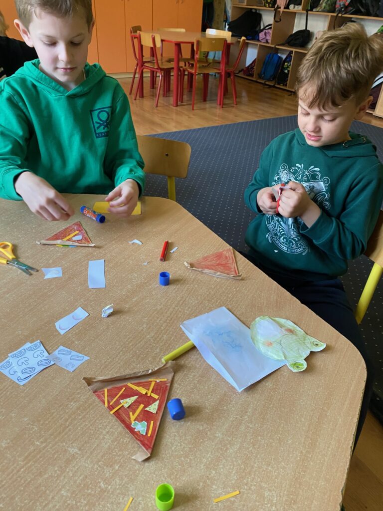 Dwóch chłopców siedzących przy stoliku przy pracy nad pizzą. Wycinają składniki do pizzy. Na stoliku kawałki pizzy i przybory plastyczne.