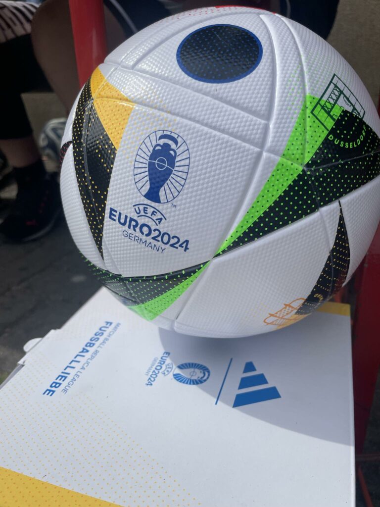 Zdjęcie przedstawia piłkę do nogi. Na białym tle piłki znajdują się kolorowe grafiki oraz napis Euro 2024