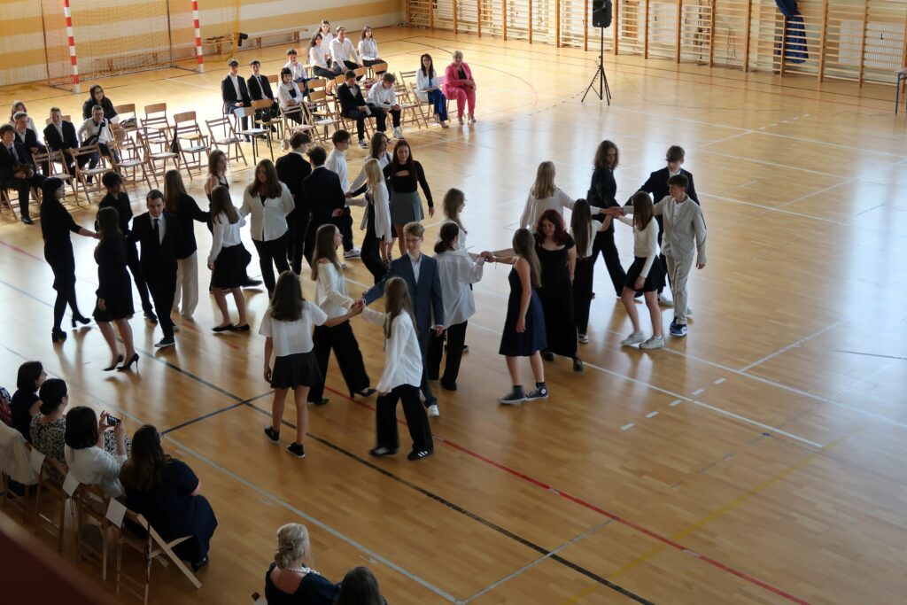 Zdjęcie przedstawia grupę chłopców i dziewcząt tańczących poloneza na sali gimnastycznej. Z prawej strony zdjęcia widać ustawione krzesła w rzędach na niektórych z nich siedzą uczniowie ubrani na galowo. Zdjęcie zrobione jest z góry.