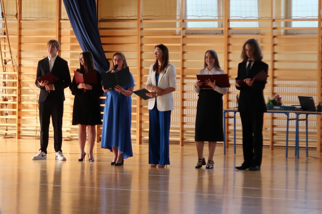 Zdjęcie przedstawia sześć osób.Dwie dziewczynki stojące z teczkami otwartymi w dłoniach. Między nimi stoją dwie kobiety. Jedna trzyma mikrofon i otwartą teczke . Ma ona na sobie niebieską sukienkę. Druga kobieta trzyma zamkniętą teczkę. Ubrana w niebieskie spodnie i białą matrynarkę.Z prawej i lewej strony stoja chłopcy ubrani w garnitury.