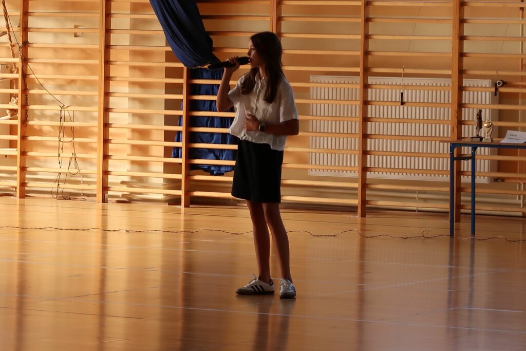 Zdjęcie przedstawia dziewczynkę ubraną na galowo trzymającą w dłoń mikrofon blisko ust. Stoi ona na tle drabinek gimnastycznych dziewczynka śpiewa.