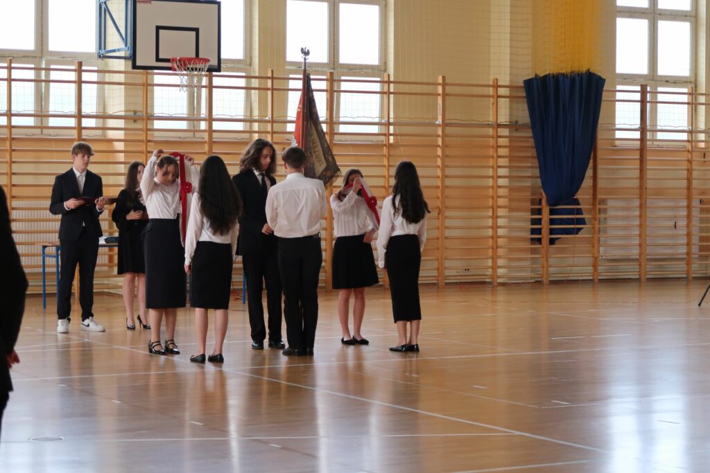 Zdjęcie przedstawia przekazanie pocztu sztandarowego. Dziewczynki ubrane na galowo ściągają biało czerwone szarfy. Chłopcy przekazują sobie sztandar. Za nimi widać dziewczynę i chłopca trzymających w ręku podkładki.