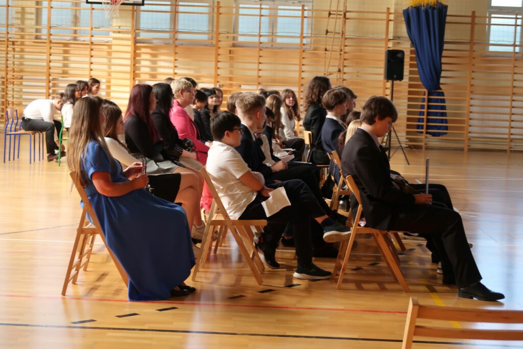 Zdjęcie przedstawia grupę chłopców i dziewcząt siedzących na krzesełkach w 3 rzędach. Pomiędzy nimi widać 2 Kobiety jedna ubrana w różowy kostium druga w niebieską sukienkę. Grupa siedzi bokiem do fotografa.