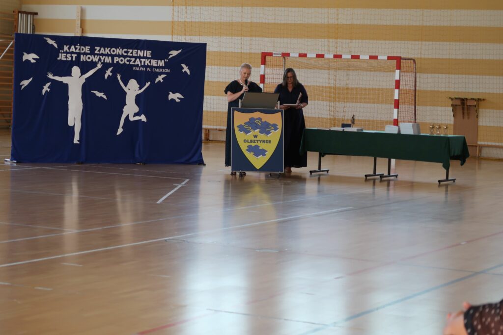 Zdjęcie przedstawia 2 Kobiety stojące za pulpitem na którym jest napis sp 7 w Olsztynie na żółtej tarczy . Jedna z kobiet trzyma mikrofon przemawia. Druga trzyma w dłoniach otwartą książkę. Kobiety stoją na tle bramki piłkarskiej. obok nich znajduje się ścianka, granatowa z napisem “każde zakończenie jest nowym początkiem” oraz grafiką chłopca i dziewczynki z wzniesionymi rękoma do góry, biegnących.
