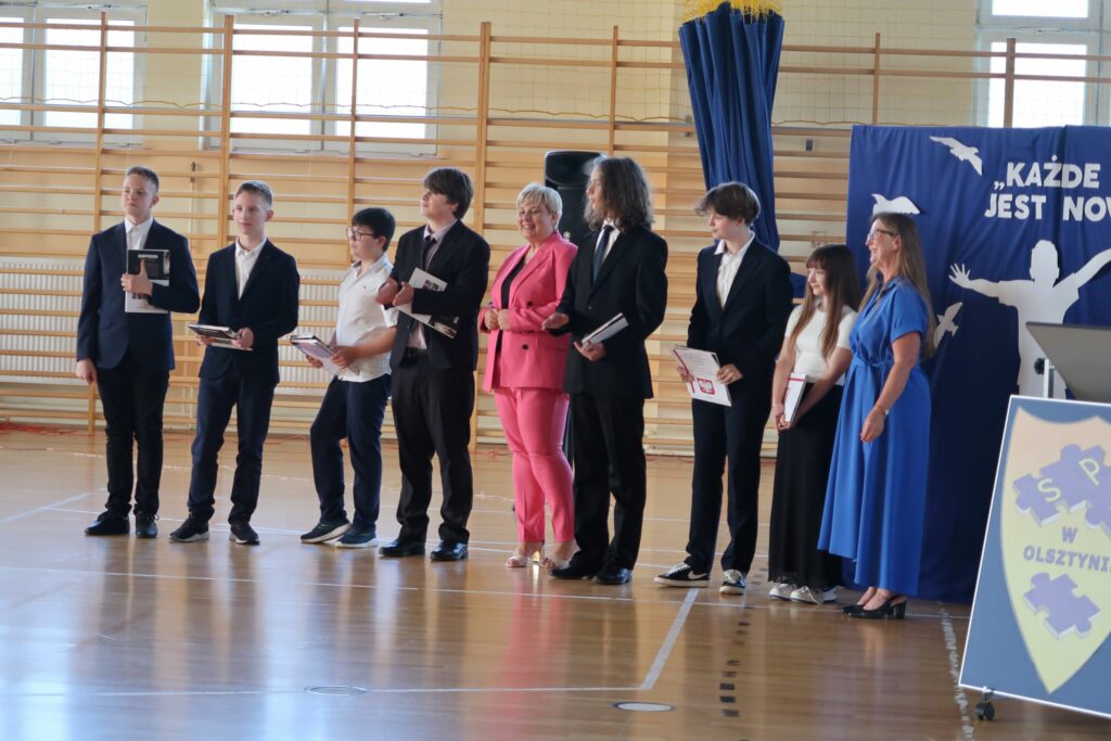 Zdjęcie przedstawia grupę chłopców i dziewcząt stojących na tle drabinek oraz granatowej ścianki. Między nimi stoją 2kobiety jedna w różowym kostiumie druga w niebieskiej sukience. Uczniowie trzymają w rękach dyplomy oraz książki.