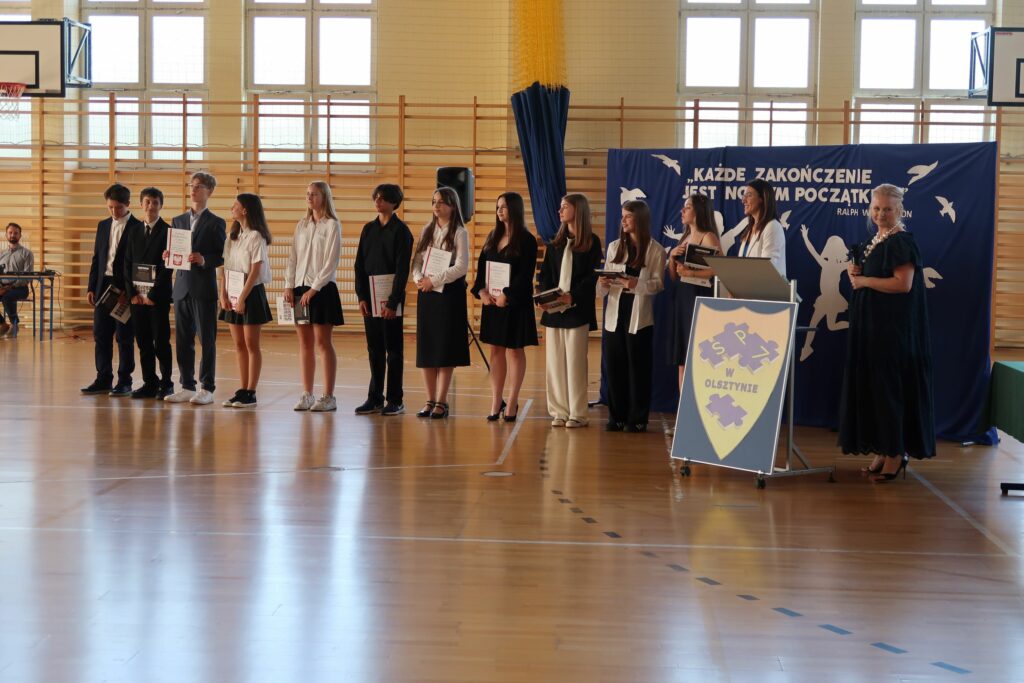 Zdjęcie przedstawia grupę chłopców i dziewcząt trzymających w ręku dyplomy i książki. Obok nich stoją 2 Kobiety jedna w granatowej sukience druga w białej marynarce. Za grupą widać granatowe tło z napisem “każdy zakończenie jest nowym początkiem”.