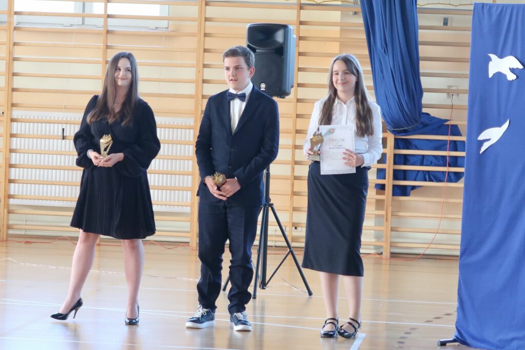 Zdjęcie przedstawia 2 dziewczynki. Pomiędzy nimi stojącego chłopca ubranego w garnitur z muszką przy szyi . Jedna z dziewcząt ubrana jest na galowo biała bluzka ciemna spódnica. Druga ma na sobie sukienkę i buty na wysokim obcasie. Wszystkie osoby trzymają w ręku statuetkę. Dziewczynka ubrana na galowo dodatkowo dyplom.