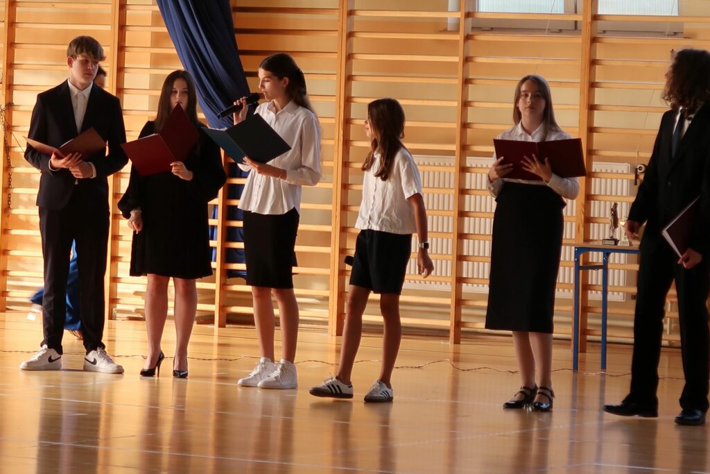 Zdjęcie przedstawia 6 osób 4 dziewczynki i 2 chłopców. Chłopcy stoją na zewnątrz grupy. Jedna z dziewczyn trzyma mikrofon oraz otwartą teczkę czyta tekst .Pozostałe słuchają, w rękach również mają otwarte teczki Pozostałe słuchają w rękach również mają otwarte teczki pozostałe słuchają w rękach również mają otwarte teczki.