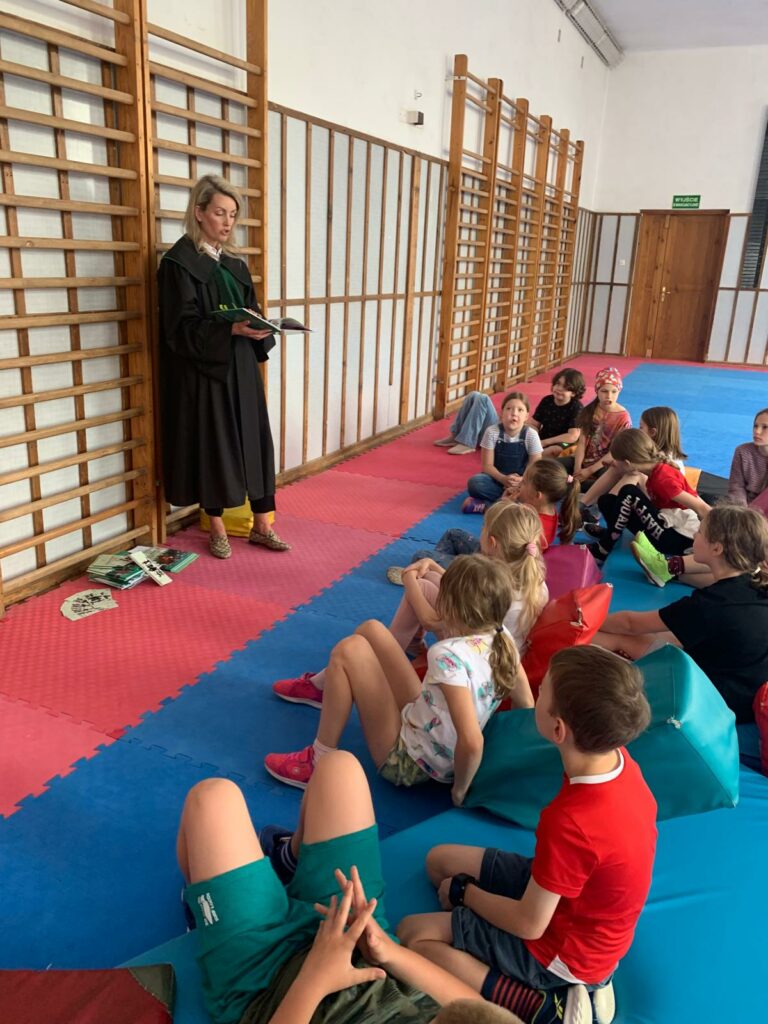 NA tle drabinek gimnastycznych stoi kobieta w todze z zielonym żabotem i trzyma w ręku książkę . Naprzeciwko kobiety grupa kilkunastu dzieci, które siedzą i leżą. Po prawej stronie kobiety leży stosik książek i zakładek. Po prawej stronie zdjęcia widać drzwi.