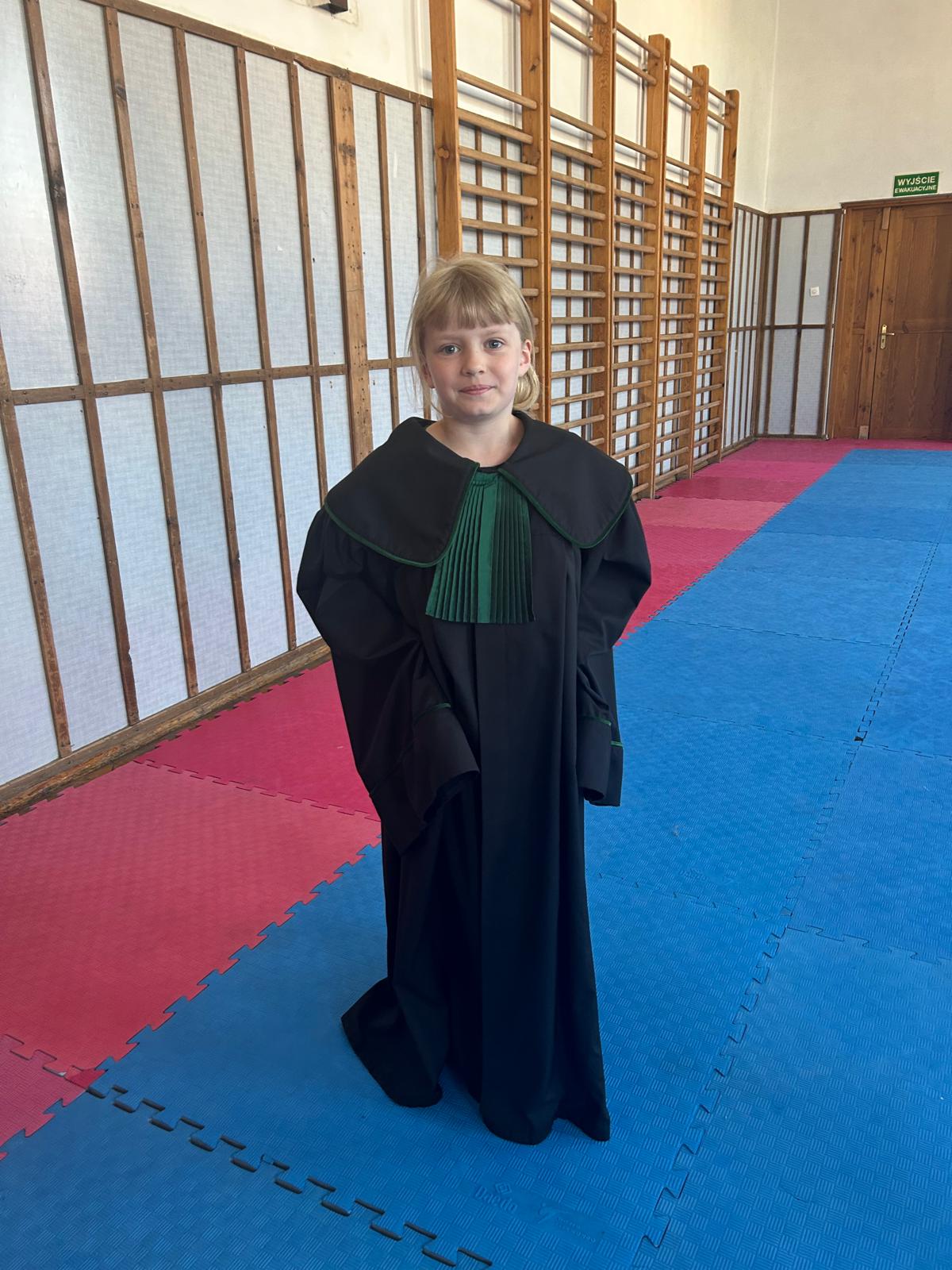 Dziewczynka w todze, w której żabot jest koloru zielonego. Ok prawej stronie dziewczynki eidać drabinki gimnastyczne.