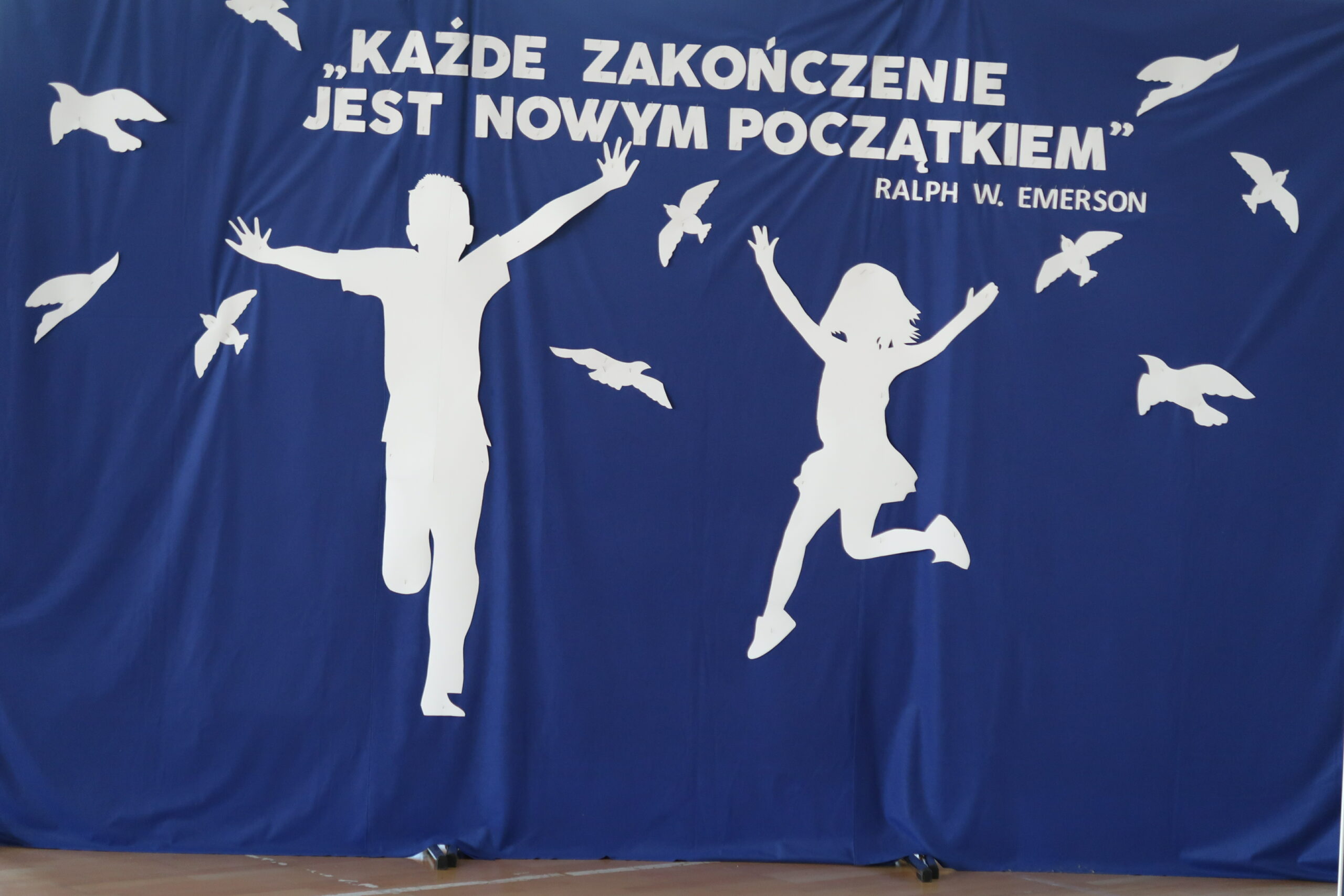 Zdjęcie przedstawia napis "Każde zakończenie jest nowym początkiem". Dzieci chłopca i dziewczynkę biegnące a wokół nich ptaki. Wszystko znajduje się na granatowym tle i jest wycięte z białego papieru.