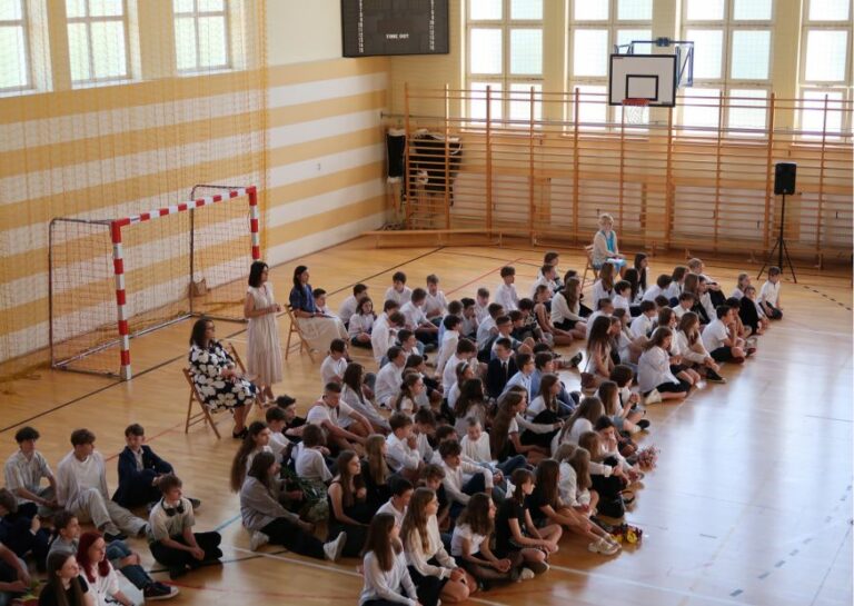 Zdjęcie zrobione z góry. Przedstawia duża grupę dzieci ubranych na galowo siedzących na podłodze na sali gimnastycznej. Między dziećmi znajdują się kobiety siedzące na krzesłach a jedna kobieta stoi.