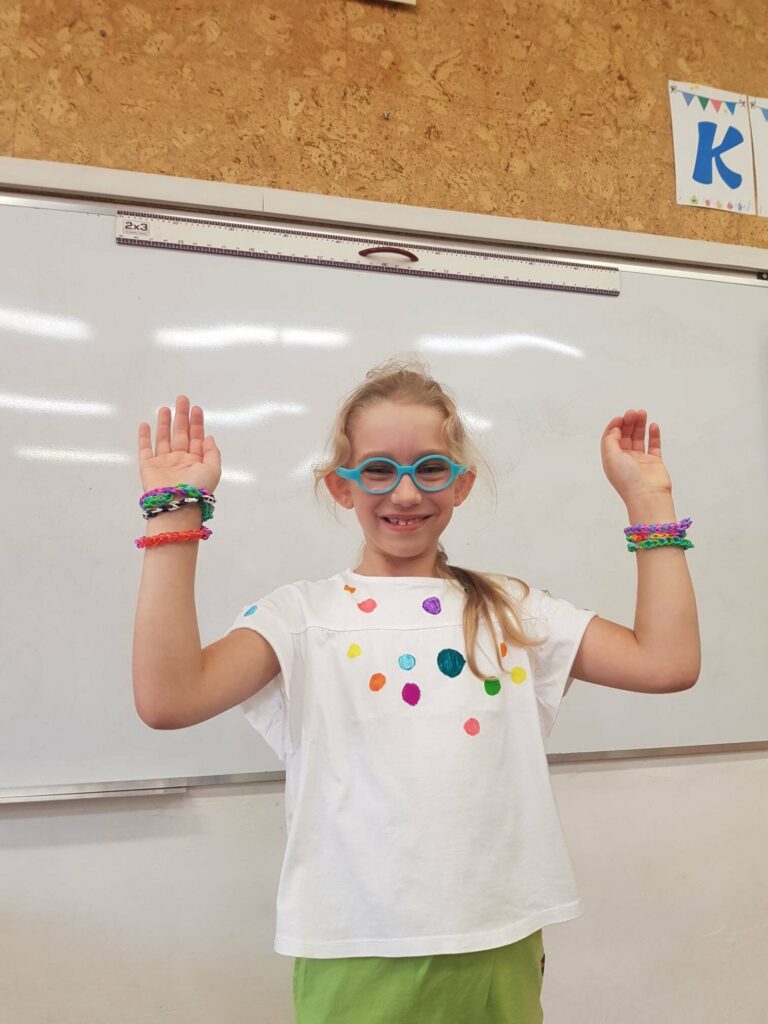 Dziewczynka stoi na tle tablicy, w klasie. Ma podniesione ręce i prezentuje bransoletki, które sama wykonała.