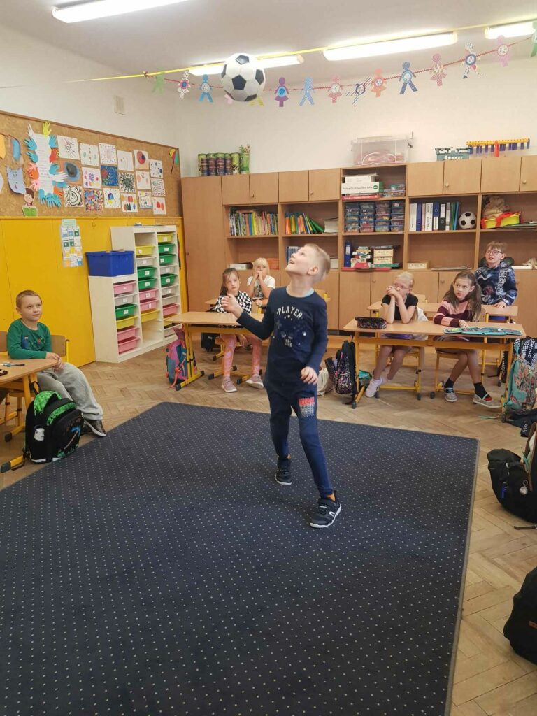 Chłopiec stoi na dywanie w klasie. Prezentuje triki z piłką. Piłka jest w górze, rzucona przez chłopca. W tle widać ławki, przy których siedzą uczniowie. Za nimi są szafy i regały.