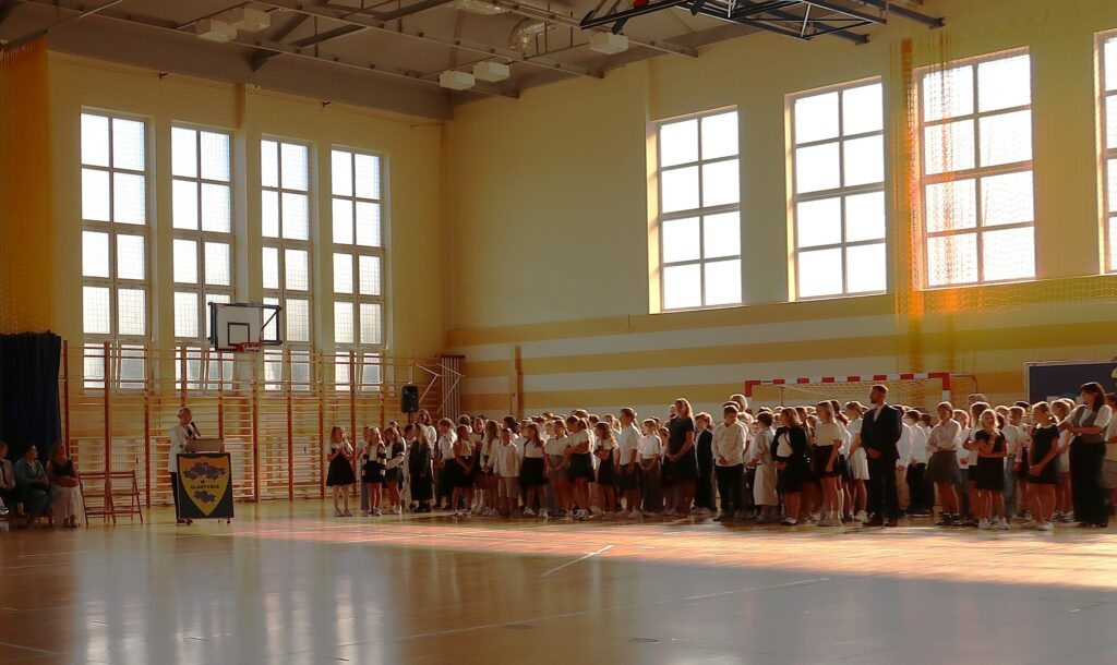 Zdjęcie przedstawia salę gimnastyczną. Na sali znajduje się duża grupa chłopców i dziewcząt ubranych na galowo. Między nimi stoją osoby dorosłe nauczyciele. Z lewej strony zdjęcia przy pulpicie stoi kobieta z mikrofonem w dłoni .