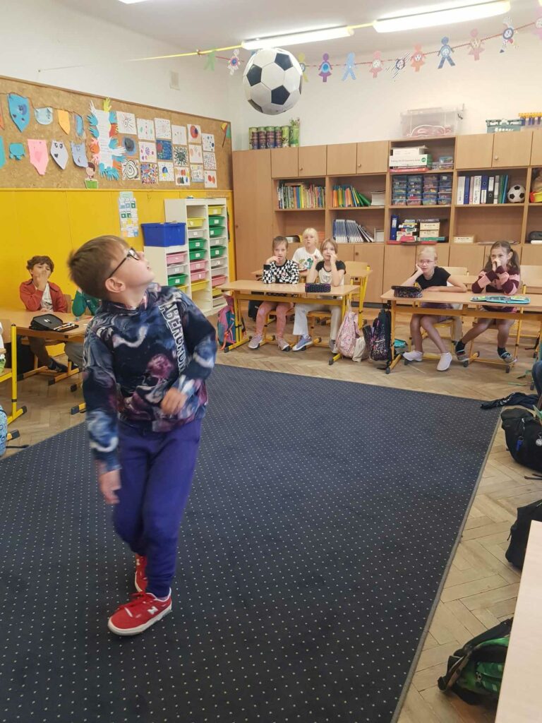 Chłopiec stoi pochylony na dywanie w klasie. Prezentuje triki z piłką. Piłka jest w górze, kopnięta przez chłopca. W tle widać ławki, przy których siedzą uczniowie. Za nimi są szafy i regały.