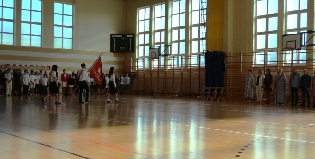 Zdjęcie przedstawia dużą salę gimnastyczną. Pod ścianę z lewej strony zdjęcia stoją uczniowie chłopcy i dziewczęta ubrani na galowo. Przed nimi widać trzy osoby. Dwie dziewczyny ubrane na galowo między nimi chłopiec również ubrany na galowo niosący sztandar szkoły. Z prawej strony zdjęcia widać stojących pod drabinkami gimnastycznymi i koszem do koszykówki mężczyzny i kobiety.