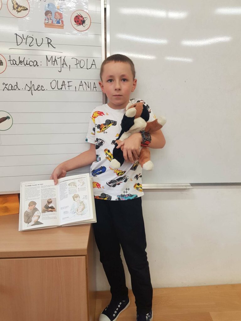 Chłopiec stoi na tle tablicy, w klasie. W jednej ręce trzyma maskotkę kotka, w drugiej- książkę o kotach.