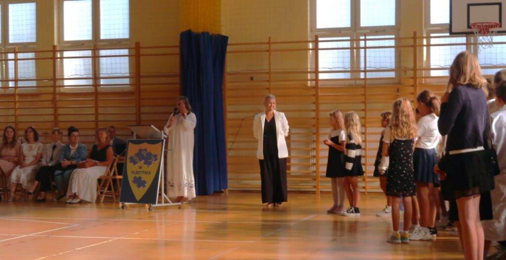 Zdjęcie przedstawia salę gimnastyczną z prawej strony zdjęcia widać grupę uczniów ubranych na galowo, stojących tyłem bądź bokiem do fotografa. Obok nich stoi kobieta ubrana w białą marynarkę i czarną sukienkę. Z twarzą zwróconą w stronę dzieci. Z jej prawej strony stoi kobieta z mikrofonem w dłoni ubrana w białą sukienkę. Przed tą kobietą znajduje się pulpit na którym jest umieszczono godło szkoły. Obok pulpitu siedzą na krzesłach kobiety.
