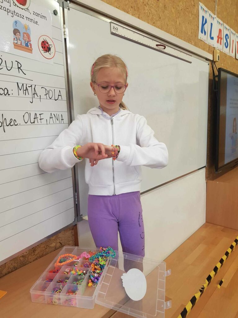 Dziewczynka stoi na tle tablicy, w klasie. Prezentuje na dłoni bransoletkę, wykonana samodzielnie. Przed nią stoi na szafce pudełko z gumkami i akcesoriami do wykonania bransoletek.