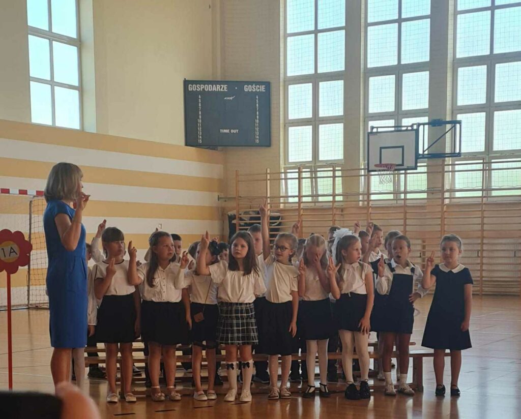 Zdjęcie przedstawia grupę dzieci w odświętnych strojach, ustawionych w sali gimnastycznej. Dzieci wznoszą palce w górę, prawdopodobnie składając ślubowanie. Obok nich stoi nauczycielka w niebieskiej sukience, prowadząca uroczystość. W tle widać tablicę wyników i kosz do koszykówki. Sala ma drewniane drabinki i duże okna.