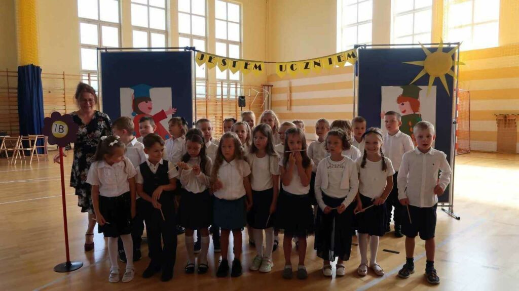 Na zdjęciu widać grupę dzieci w wieku szkolnym, stojących na sali gimnastycznej. Są one ubrane elegancko, bo uczestniczą w ważnym wydarzeniu szkolnym, pasowaniu na ucznia. Nauczycielka, stojąca z lewej strony, trzyma tabliczkę z napisem "1B". W tle widoczna jest dekoracja z napisem "JESTEM UCZNIEM" oraz symbolem słońca, co dodatkowo podkreśla uroczysty charakter wydarzenia. Jest to na sali gimnastycznej. W tle okna i drabinki.