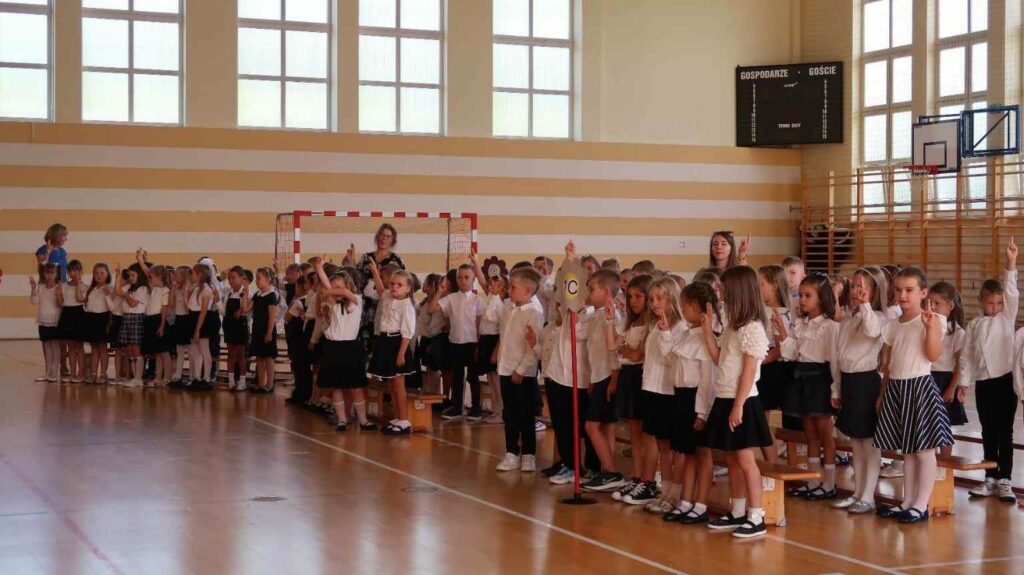 Na zdjęciu znajduje się grupa dzieci w sali gimnastycznej, podczas ceremonii szkolnej. Uczniowie są ubrani w eleganckie białe koszule oraz ciemne spódnice lub spodnie. Dzieci stoją w rzędach, a większość z nich unosi prawą rękę, bo składały przysięgę. W tle widać tablicę wyników sportowych oraz bramkę do gry w piłkę ręczną, co sugeruje, że wydarzenie odbywa się w hali sportowej. Przy dzieciach znajdują się nauczycielki, nadzorujące ceremonię. Pomieszczenie jest dobrze oświetlone, dzięki dużym oknom, które wpuszczają sporo naturalnego światła.