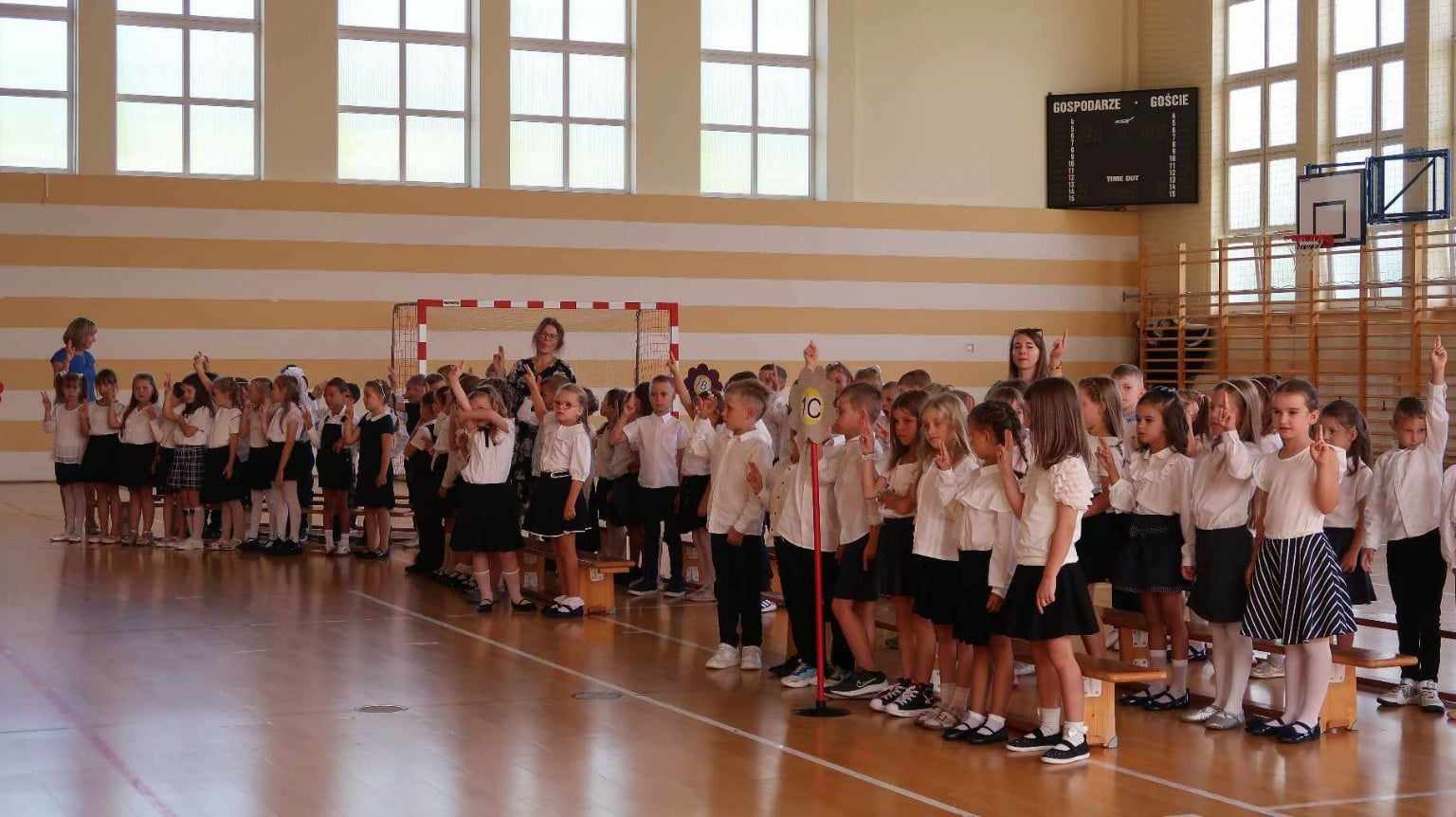 Na zdjęciu znajduje się grupa dzieci w sali gimnastycznej, podczas ceremonii szkolnej. Uczniowie są ubrani w eleganckie białe koszule oraz ciemne spódnice lub spodnie. Dzieci stoją w rzędach, a większość z nich unosi prawą rękę, bo składały przysięgę. W tle widać tablicę wyników sportowych oraz bramkę do gry w piłkę ręczną, co sugeruje, że wydarzenie odbywa się w hali sportowej. Przy dzieciach znajdują się nauczycielki, nadzorujące ceremonię. Pomieszczenie jest dobrze oświetlone, dzięki dużym oknom, które wpuszczają sporo naturalnego światła.