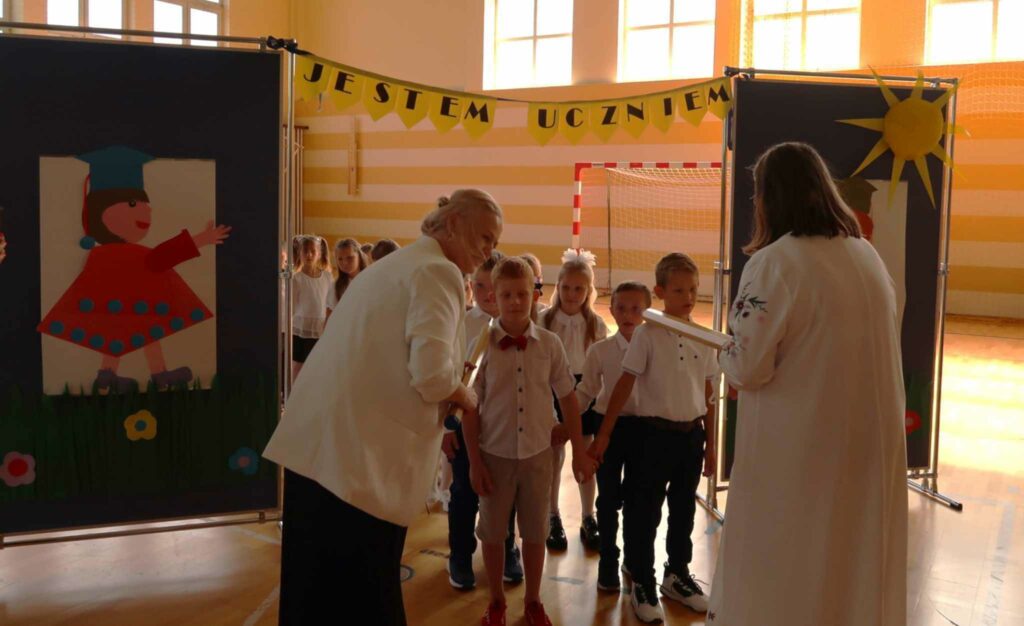 Na zdjęciu widać grupę dzieci, które stoją w rzędzie podczas uroczystości szkolnej. Dzieci są ubrane w odświętne stroje – białe koszule, a jedno z nich ma na sobie muszkę. Panie dyrektorki uczestniczą w ceremonii, jedna z nich trzyma duży ołówek. W tle widać dekoracje szkolne, w tym kolorowy napis "JESTEM UCZNIEM" oraz obrazek przedstawiający dziecko w birecie. Całość odbywa się w sali gimnastycznej, a atmosfera jest oficjalna i uroczysta.