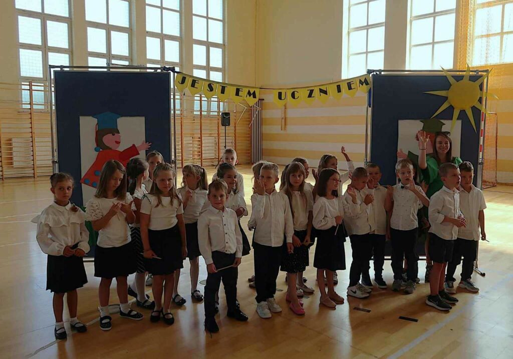 Na zdjęciu widać grupę dzieci w wieku szkolnym, stojących na sali gimnastycznej. Są one ubrane elegancko, bo uczestniczą w ważnym wydarzeniu szkolnym, pasowaniu na ucznia. Nauczycielka, stojąca z lewej strony, trzyma tabliczkę z napisem "1B". W tle widoczna jest dekoracja z napisem "JESTEM UCZNIEM" oraz symbolem słońca, co dodatkowo podkreśla uroczysty charakter wydarzenia. Jest to na sali gimnastycznej. W tle okna i drabinki.