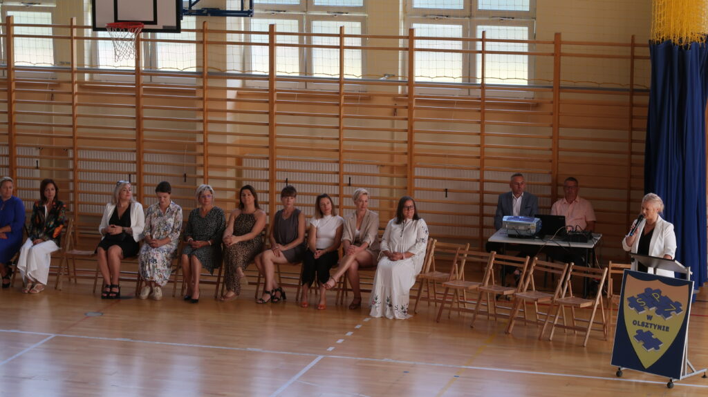 Zdjęcie przedstawia 10 kobiet siedzących na krzesełkach pod drabinkami gimnastycznymi na sali gimnastycznej. Obok nich znajdują się puste krzesła. Przy nich stoi kobieta ubrana w białą marynarkę trzymająca w jednej dłoni mikrofon a drugą rękę opiera o pulpit stojący przed nią. Za pustymi krzesłami widać dwóch mężczyzn siedzących przy stoliku, na którym znajduje się sprzęt muzyczny.