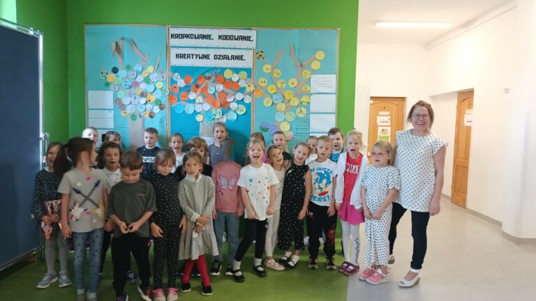 Na pierwszym planie są uśmiechnięte dzieci – dziewczynki i chłopcy. Dzieci ubrane są w kolorowe ubrania z motywem kropek. W tle widać tablicę z niebieskim tłem i papierowymi drzewami, na których przyczepione są kolorowe kółeczka z narysowanymi przez dzieci talentami.