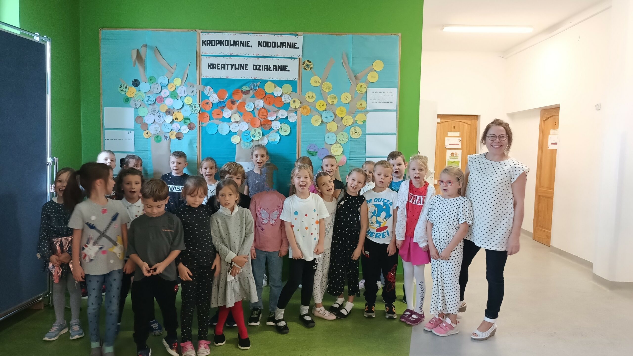 Na pierwszym planie są uśmiechnięte dzieci – dziewczynki i chłopcy. Dzieci ubrane są w kolorowe ubrania z motywem kropek. W tle widać tablicę z niebieskim tłem i papierowymi drzewami, na których przyczepione są kolorowe kółeczka z narysowanymi przez dzieci talentami.