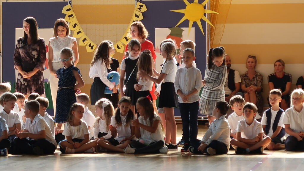 Zdjęcie przedstawia grupę chłopców i dziewcząt ubranych na galowo. Znaczna część dzieci siedzi po turecku. Między nimi znajdują się stojące dzieci rozmawiające ze sobą, odwrócone do fotografa w różne strony. Między dziećmi stoją trzy kobiety jedna ubrana na czerwono, druga ubrana na biało, trzecia ma ciemną sukienkę. Za grupą tych osób znajdują się plansze między którymi wisi napis “Jestem uczniem “obok znajduje się papierowe słońce przypięte do tablicy. Przy tych planszach siedzą pod ścianą na ławce trzy osoby kobiety i mężczyzna.