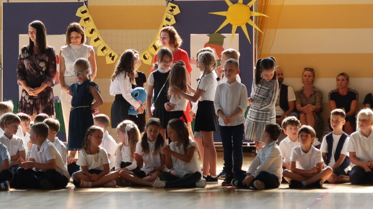 Zdjęcie przedstawia grupę chłopców i dziewcząt ubranych na galowo. Znaczna część dzieci siedzi po turecku. Między nimi znajdują się stojące dzieci rozmawiające ze sobą, odwrócone do fotografa w różne strony. Między dziećmi stoją trzy kobiety jedna ubrana na czerwono, druga ubrana na biało, trzecia ma ciemną sukienkę. Za grupą tych osób znajdują się plansze między którymi wisi napis “Jestem uczniem “obok znajduje się papierowe słońce przypięte do tablicy. Przy tych planszach siedzą pod ścianą na ławce trzy osoby kobiety i mężczyzna.