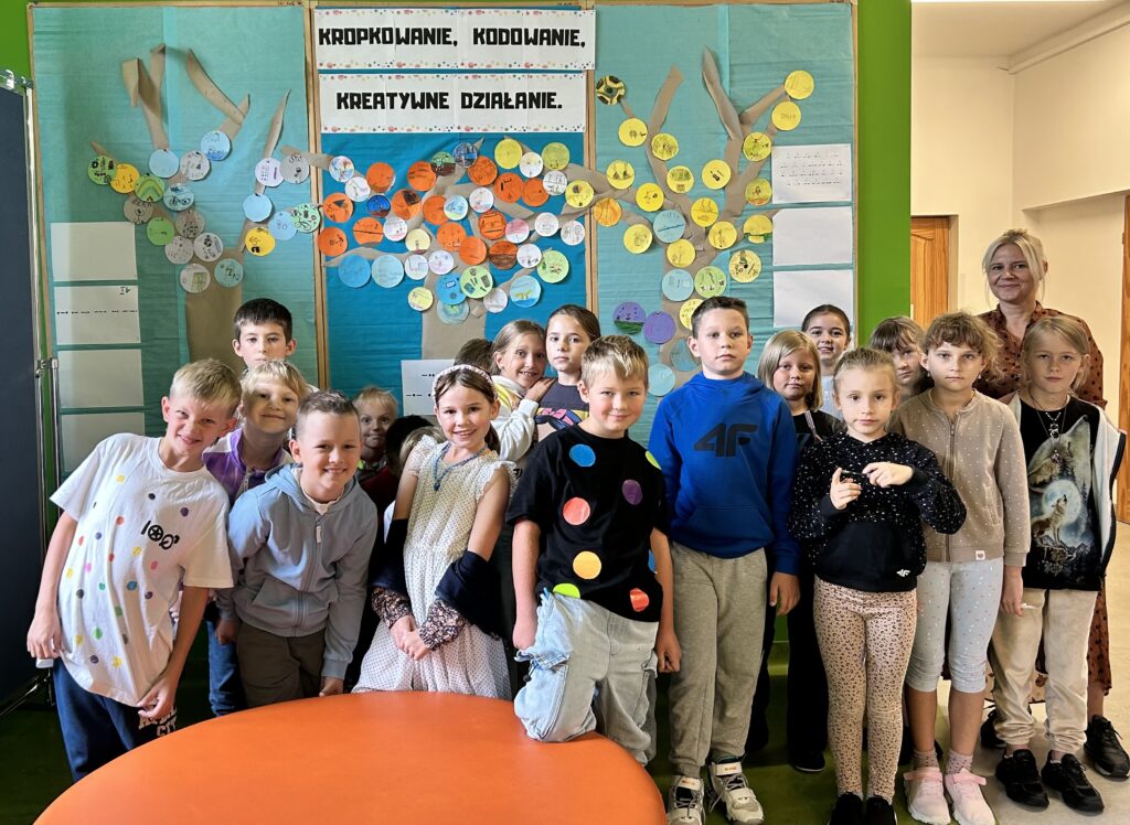 Na zdjęciu na pierwszym planie są uśmiechnięte dzieci – dziewczynki i chłopcy. Większość z nich włożyła kolorowe ubrania z motywem kropek. W tle widać tablicę z niebieskim tłem i papierowymi drzewami, na których przyczepione są kolorowe kółeczka z narysowanymi przez dzieci talentami.