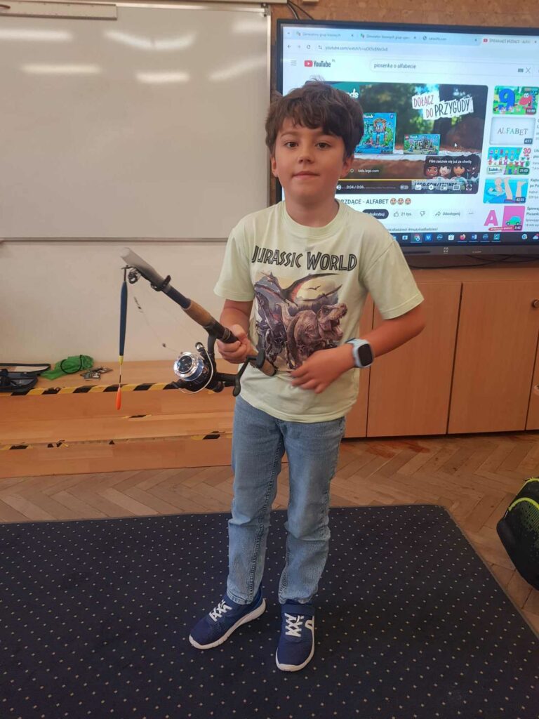 Chłopiec stoi na dywanie w klasie. W ręku trzyma wędkę. Za nim widać białą tablicę i ekran.