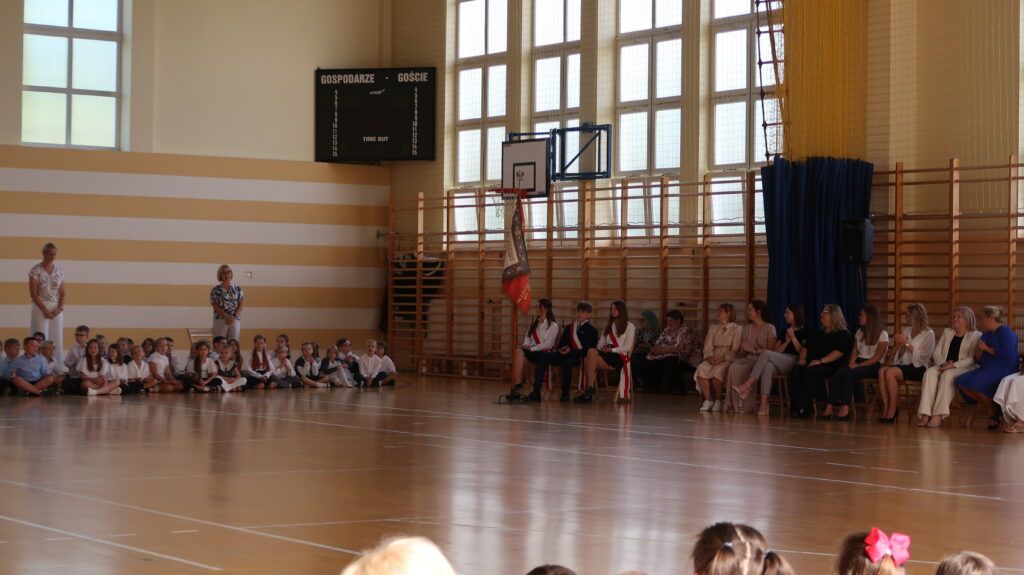 Zdjęcie przedstawia salę gimnastyczną, na której po lewej stronie zdjęcia znajdują się chłopcy i dziewczęta w dużej grupie. Siedzą oni na podłodze ubrani są na galowo. Między dziećmi stoją dwie kobiety ubrane w jasne spodnie i jasne bluzki. Obok nich widać sztandar szkoły i siedzących przy nim poczet sztandarowy. Za pocztem na krzesłach siedzą kobiety, za nimi widać drabinki gimnastyczne kosz do koszykówki duże okna.