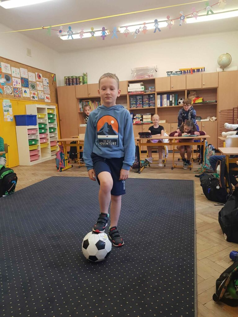 Chłopiec stoi na dywanie w klasie. Przed nim leży piłka nożna, chłopiec opiera o nią jedną nogę. W tle widać ławki, przy których siedzą uczniowie. Za nimi są szafy i regały.