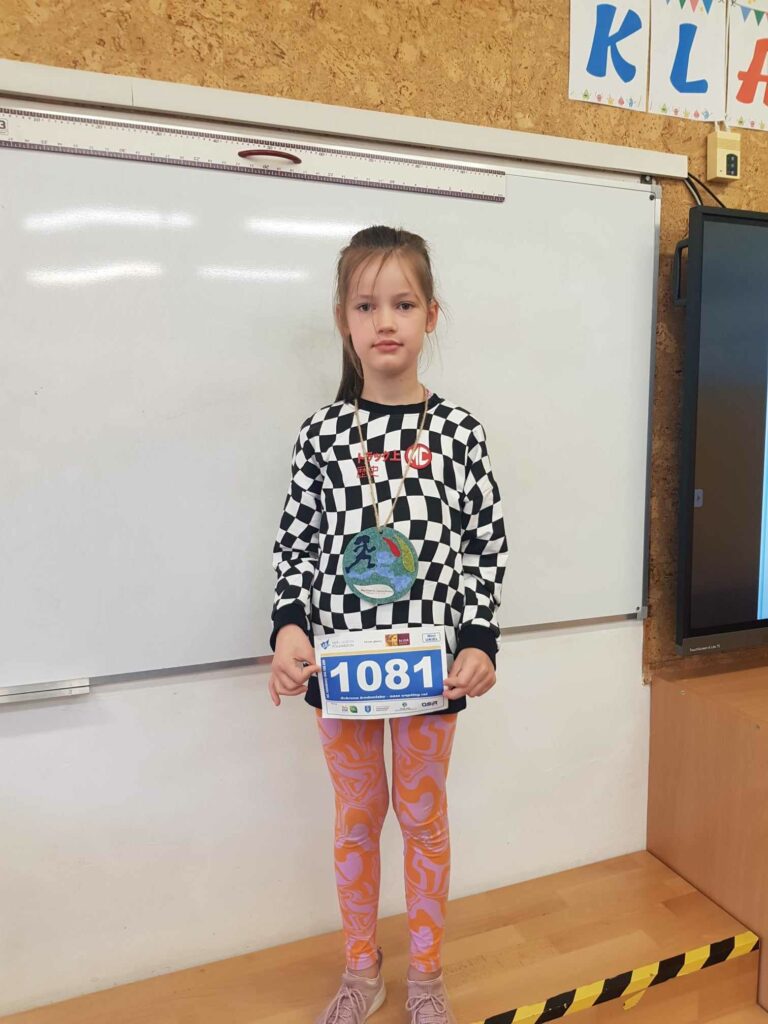 Dziewczynka stoi w klasie, na tle tablicy. W rękach trzyma numer startowy z zawodów, na szyi wisi medal. Za nią jest biała tablica.