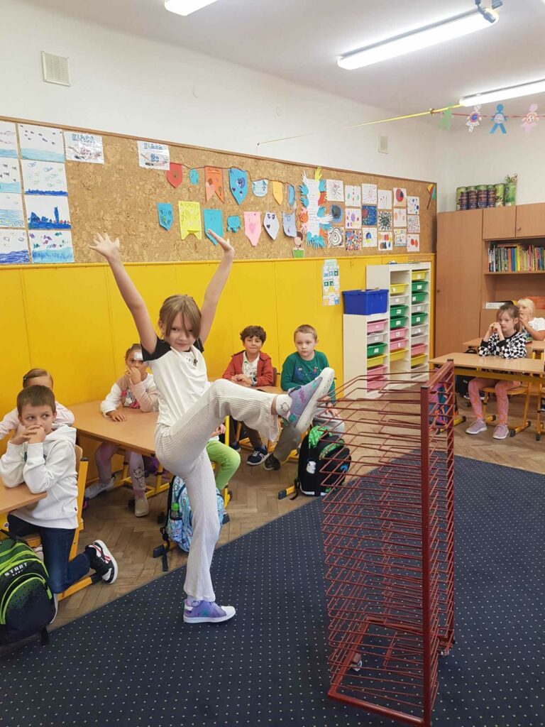 Dziewczynka stoi na dywanie w klasie. ma wysoko podniesioną nogę, opartą o stojak na prace plastyczne. Ręce podniosła wysoko. Za nią są ławki, przy których siedzą uczniowie oraz tablica z pracami plastycznymi.
