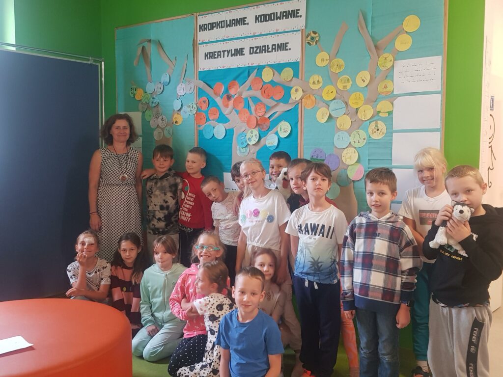 Na zdjęciu na pierwszym planie są uśmiechnięte dzieci – dziewczynki i chłopcy. Większość z nich włożyła kolorowe ubrania z motywem kropek. W tle widać tablicę z niebieskim tłem i papierowymi drzewami, na których przyczepione są kolorowe kółeczka z narysowanymi przez dzieci talentami.