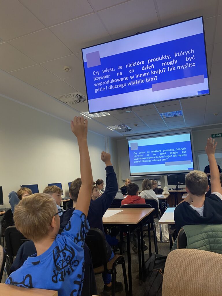 Na zdjęciu grupa dzieci siedzących w ławkach zdjęcie zrobione jest od tyłu widać plecy dzieci dzieci trzymają ręce w górze ponieważ zgłaszają się do odpowiedzi na pytanie które widnieje na ekranie prze nimi. Na zdjęciu widać dwa ekrany jeden dalej a drugi bliżej aby te dzieci siedzące dalej również mogły odczytać informacje z ekranu. Na zdjęciu na pierwszym planie widać chłopca w niebieskiej koszulce na krótki rękaw w jasnych włosach i okularkach trzyma on rękę w górze troszkę dalej z ławce witać siedzącego chłopca z granatowej ciemnej bluzie również trzyma rękę w górze a z prawej strony chłopca siedzi inny chłopiec z jasnymi włoskami które również trzyma rękę w górze na siedzeniu ma zawieszono oparcie zieloną kamizelkę.