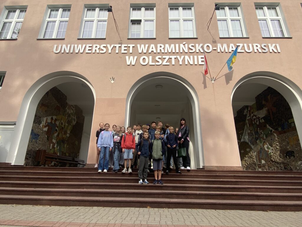 Na zdjęciu Grupka dzieci stoi na schodach przed Rektoratem uniwersytetu warmińsko-mazurskiego. Stoją one pod napisem uniwersytet warmińsko mazurski w Olsztynie. Dzieci stoją na tle budynku który posiada trzy wejścia w kształcie łuku. Budynek jest koloru rudawego na budynku wiszą dwie flagi, Jedna jest to flaga biało czerwona druga niebieska flaga uniwersytetu warmińsko-mazurskiego.