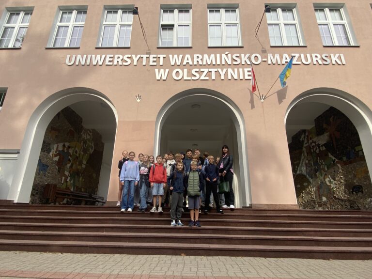 Na zdjęciu Grupka dzieci stoi na schodach przed Rektoratem uniwersytetu warmińsko-mazurskiego. Stoją one pod napisem uniwersytet warmińsko mazurski w Olsztynie. Dzieci stoją na tle budynku który posiada trzy wejścia w kształcie łuku. Budynek jest koloru rudawego na budynku wiszą dwie flagi, Jedna jest to flaga biało czerwona druga niebieska flaga uniwersytetu warmińsko-mazurskiego.